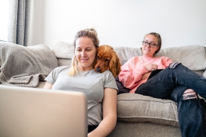 A low angle view of a female couple sitting in their living room together while working from home during the COVID-19 pandemic. One woman is using a laptop on the floor while the other is using a digital tablet on the sofa and looking at her fiancée on the floor. Their dog is sitting on the sofa with his head on the shoulder of the woman on the floor.